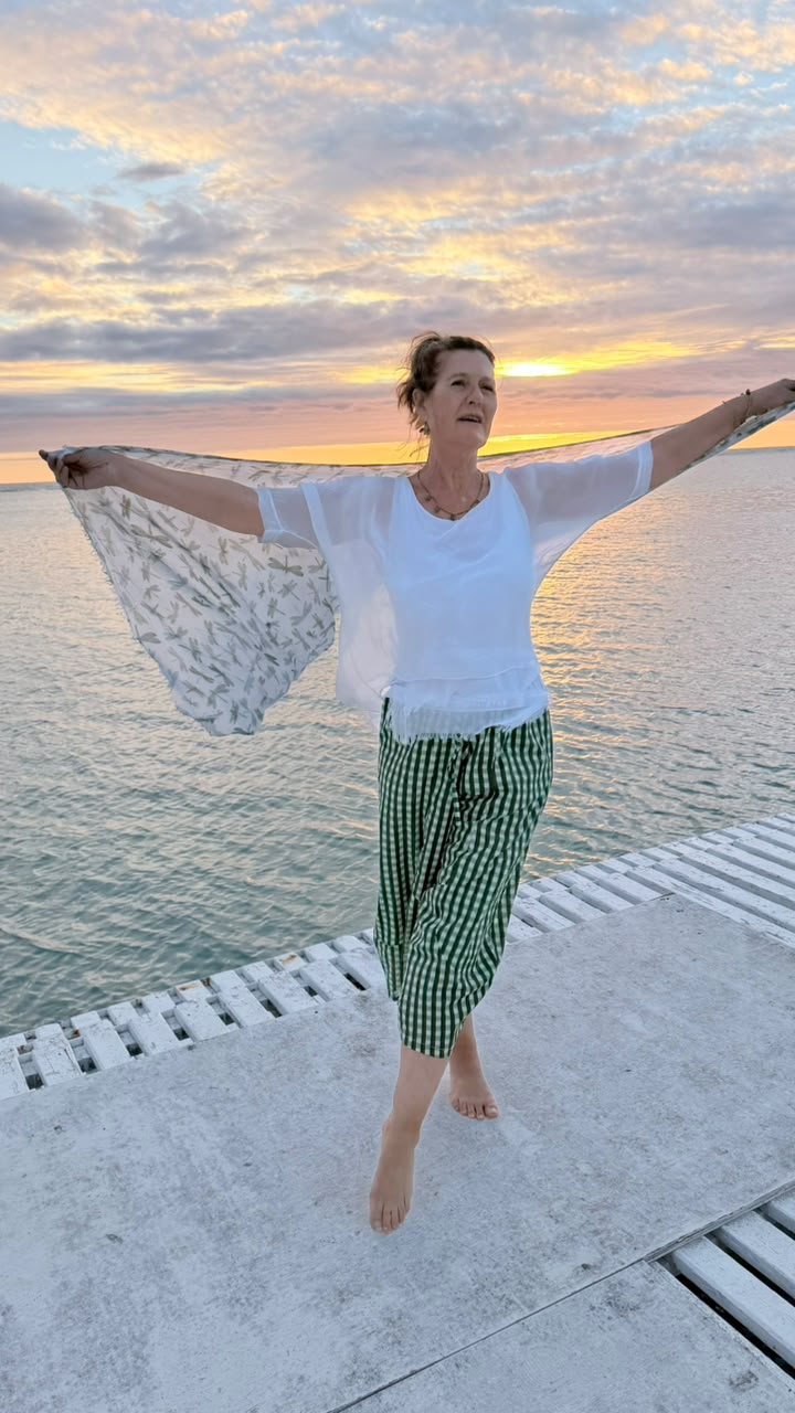 A woman stands barefoot on a concrete pier with her arms outstretched, wearing a white short-sleeve top, green and white checkered wrap-style pants, and holding a sheer white patterned scarf against a sunset over calm water.