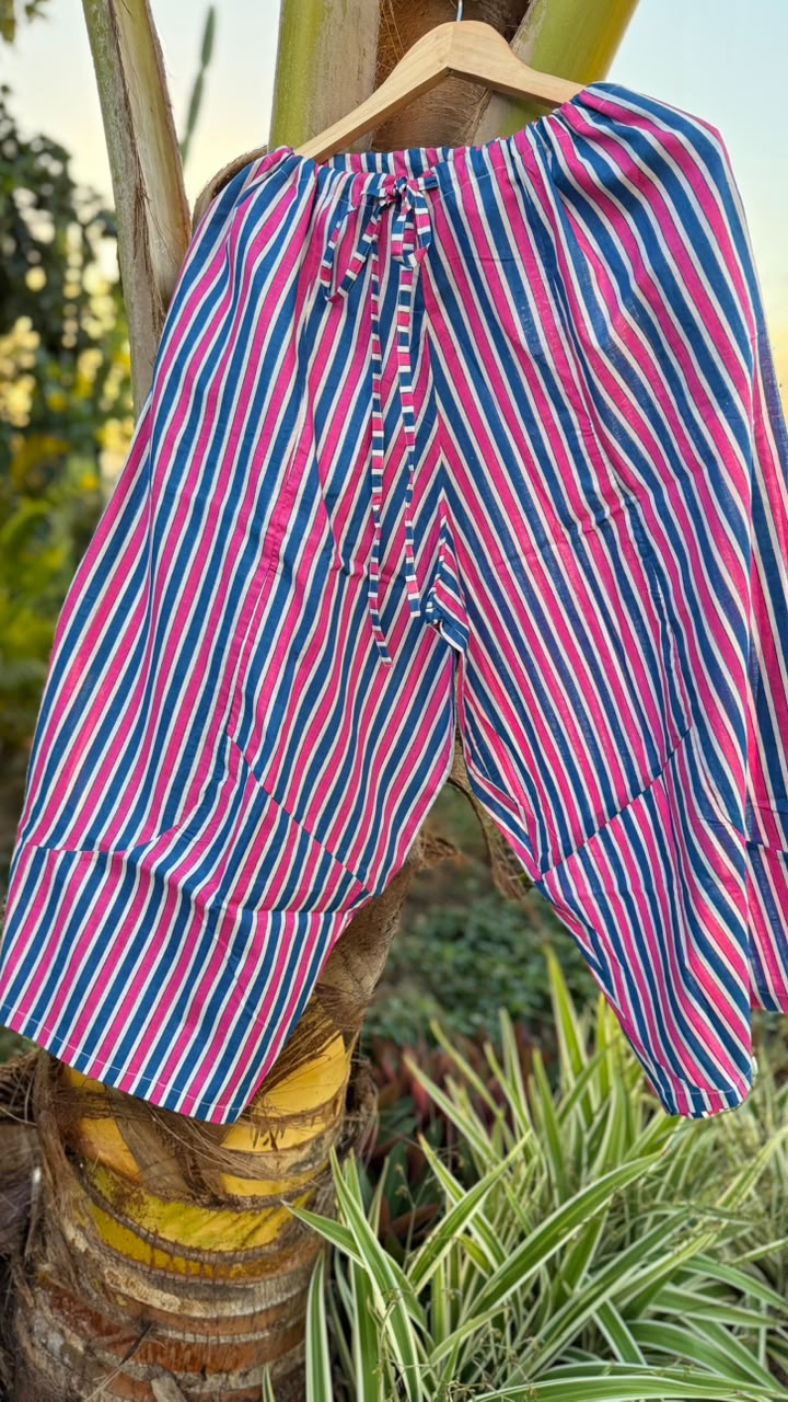 Pink, blue, and white striped cotton pants hanging on a wooden hanger outdoors against a tropical plant background.