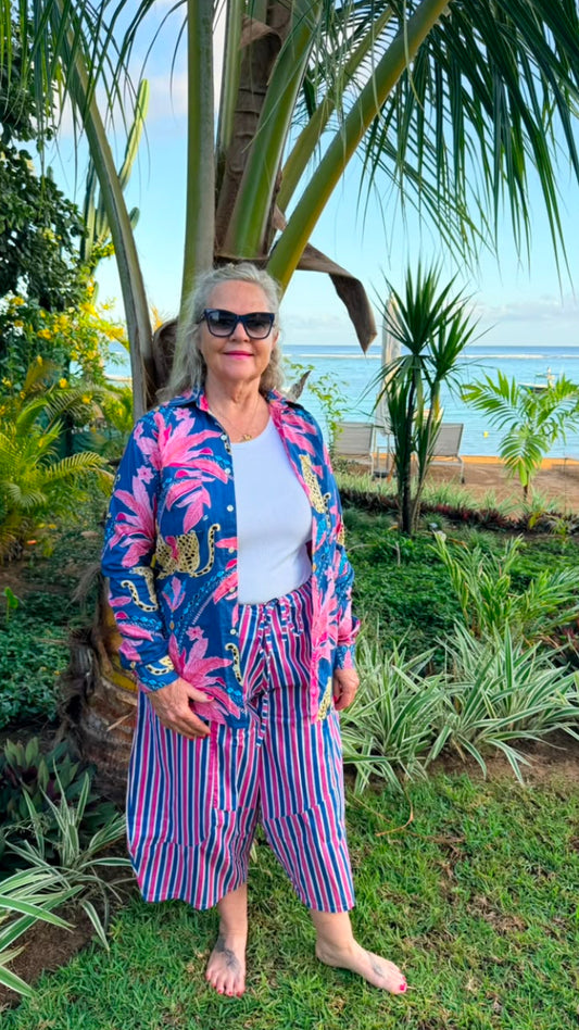 A woman stands barefoot in a tropical garden setting, wearing a vibrant blue and pink floral patterned jacket over a white top, paired with matching pink, purple, and blue striped pants.