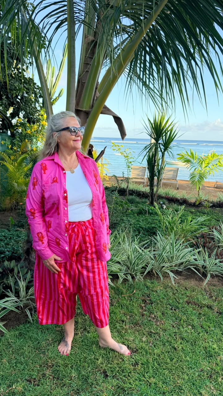 A woman wearing a vibrant pink floral button-up shirt and matching pink striped pants stands barefoot on a grassy lawn with a tropical beach and palm trees in the background.