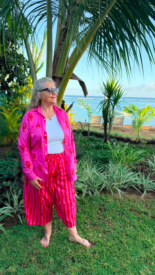 A woman wearing a vibrant pink floral button-up shirt and matching pink striped pants stands barefoot on a grassy lawn with a tropical beach and palm trees in the background.