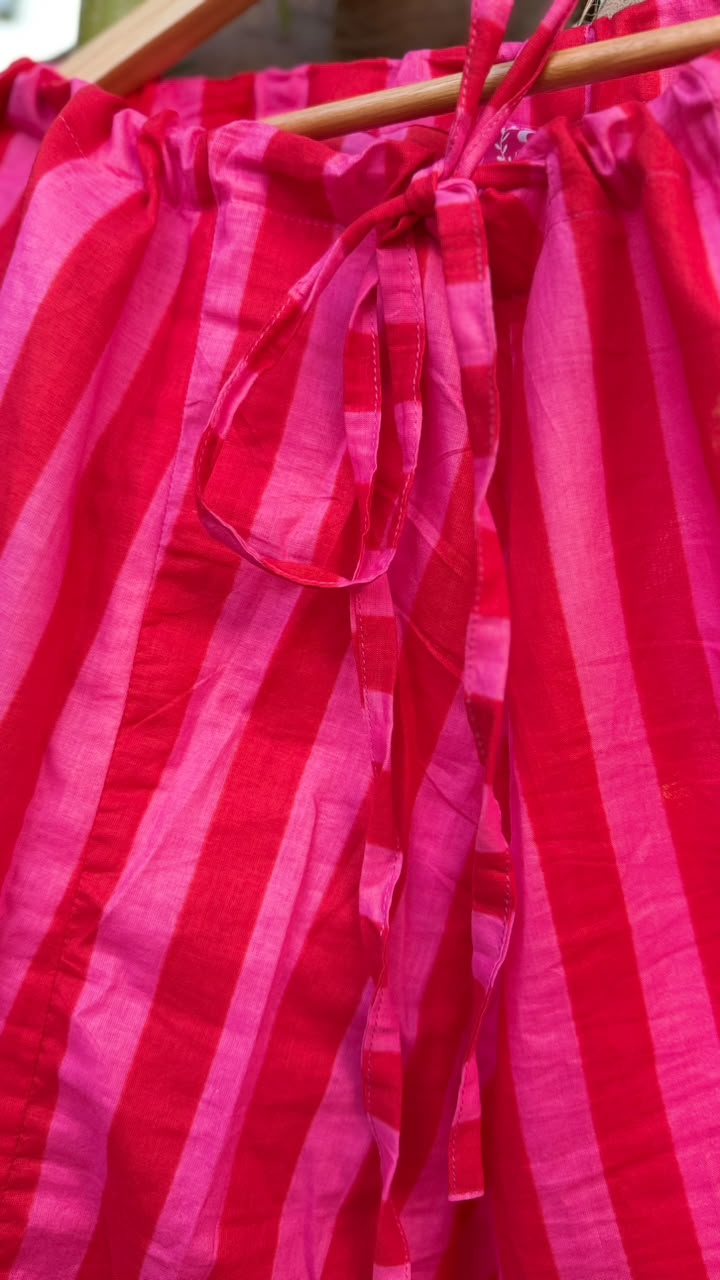 A close-up of a bright pink and red striped fabric garment hanging on a wooden hanger, with a drawstring tie at the waist.