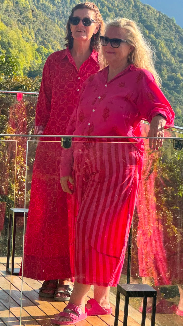 Two women wearing matching bright pink and red patterned lounge sets or sarongs stand on a wooden deck with a glass railing, with a lush green mountain backdrop.