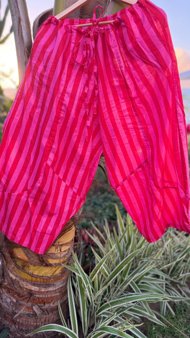Bright pink and red vertically striped cotton harem pants hanging on a wooden hanger outdoors against a palm tree trunk and green plants.