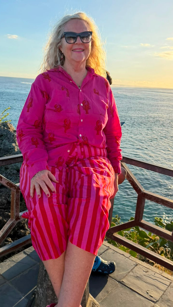 A woman wearing a bright pink matching lounge set with red patterns on a hooded long-sleeve top and striped shorts, standing outdoors by the ocean at sunset.