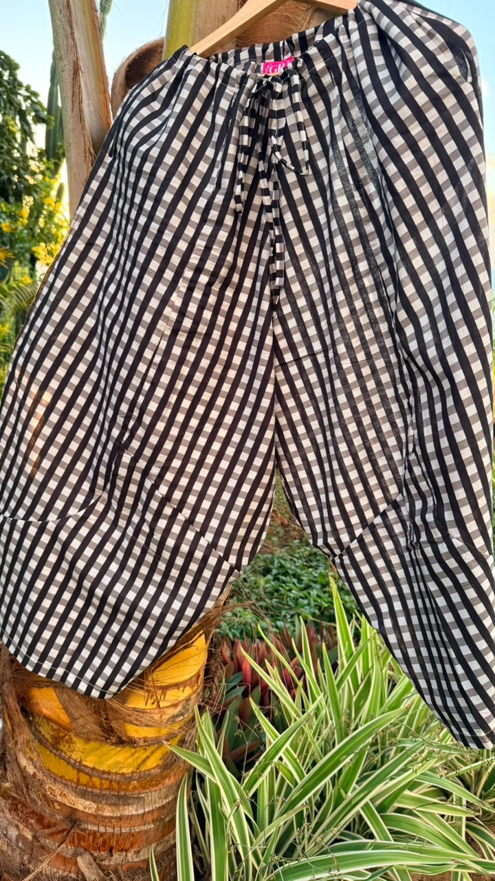 Black and white gingham checkered pants hanging on a wooden hanger outdoors in front of a tree trunk and green plants.