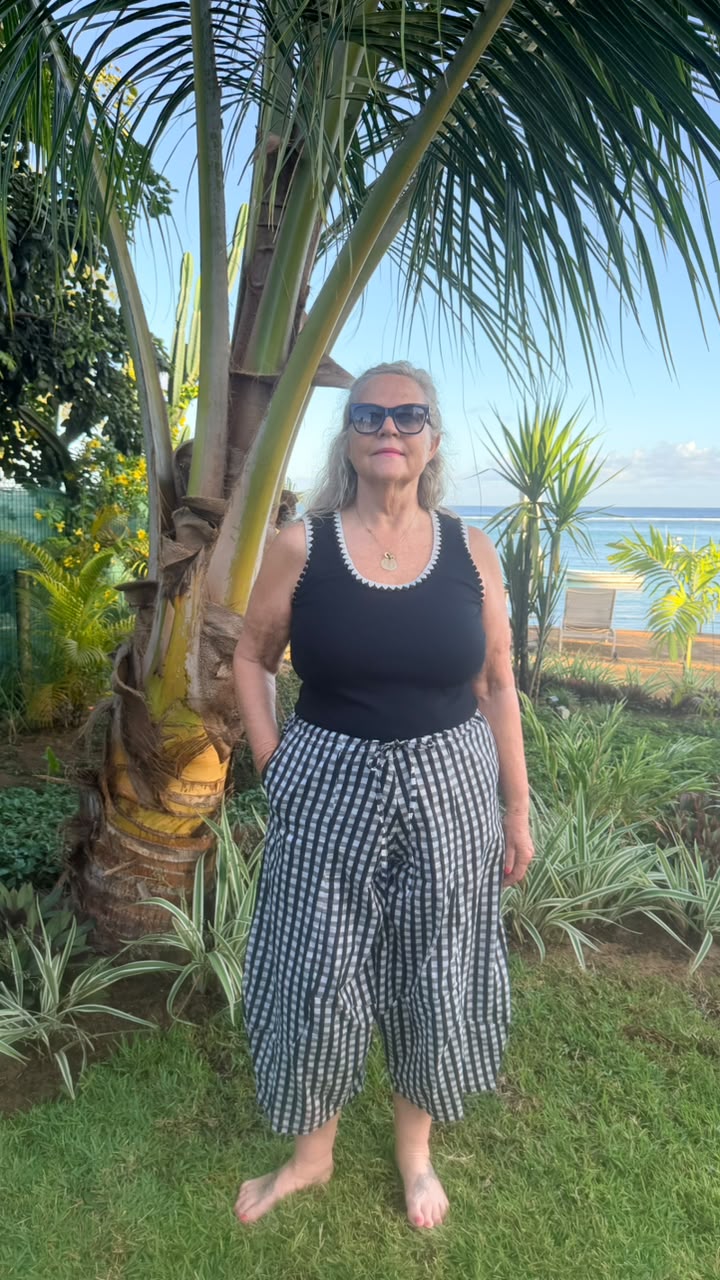 A woman standing outdoors in front of a palm tree wearing a black tank top and black-and-white checkered wide-leg pants, with ocean and lounge chairs visible in the background.