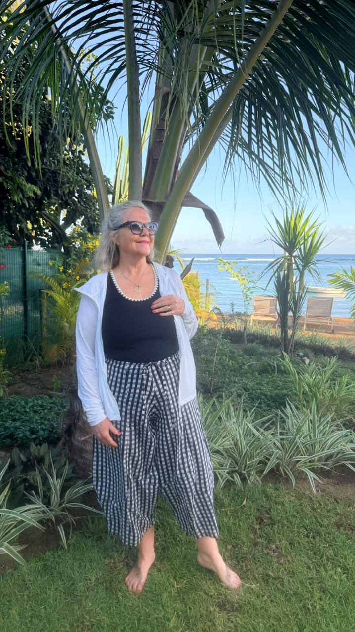 A woman stands barefoot on a grassy lawn outdoors, wearing a black tank top, a white zip-front cardigan, and black-and-white gingham cropped pants. Palm trees and a garden are behind her, with ocean chairs and water visible in the distance.