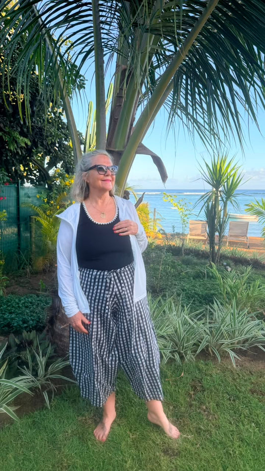 A woman stands barefoot on a grassy lawn outdoors, wearing a black tank top, a white zip-front cardigan, and black-and-white gingham cropped pants. Palm trees and a garden are behind her, with ocean chairs and water visible in the distance.