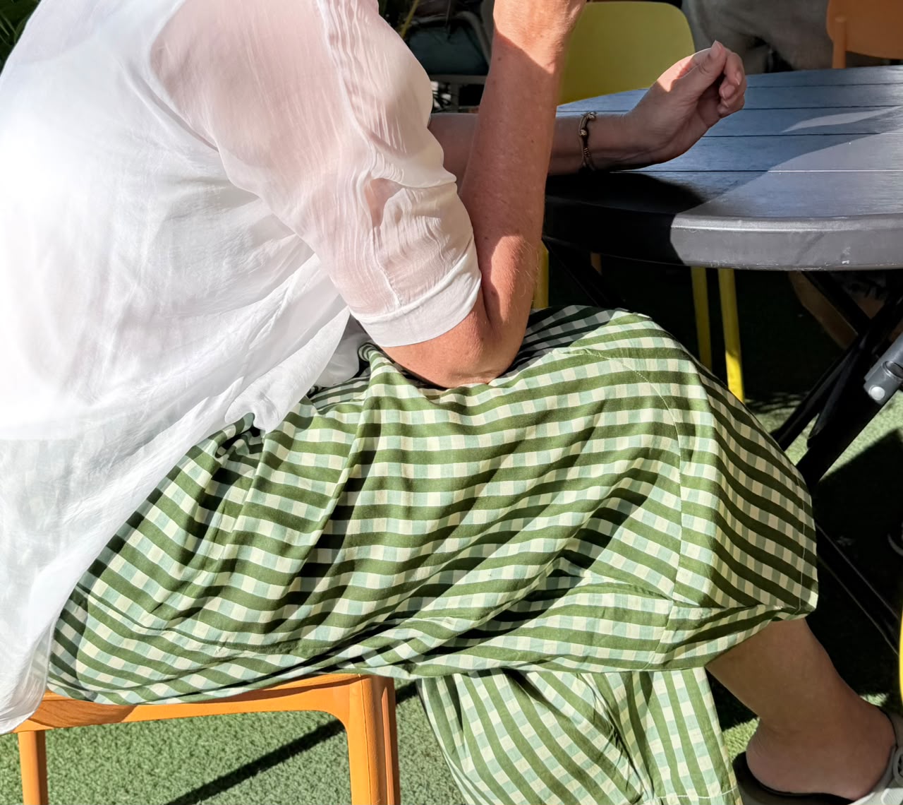 A person seated at an outdoor table wearing a green and white gingham skirt and a sheer white top, photographed from the side in natural sunlight.