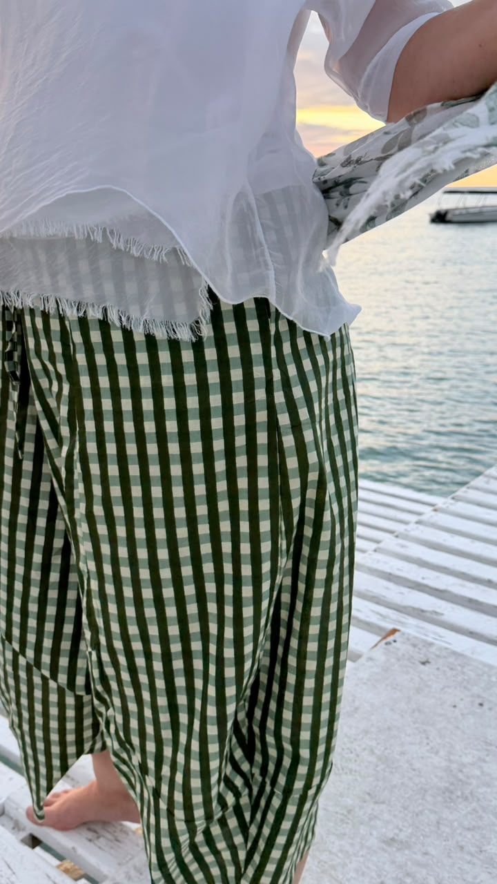 A person standing on a white wooden dock by the water at sunset, wearing green and white gingham checkered pants and a white sheer top.