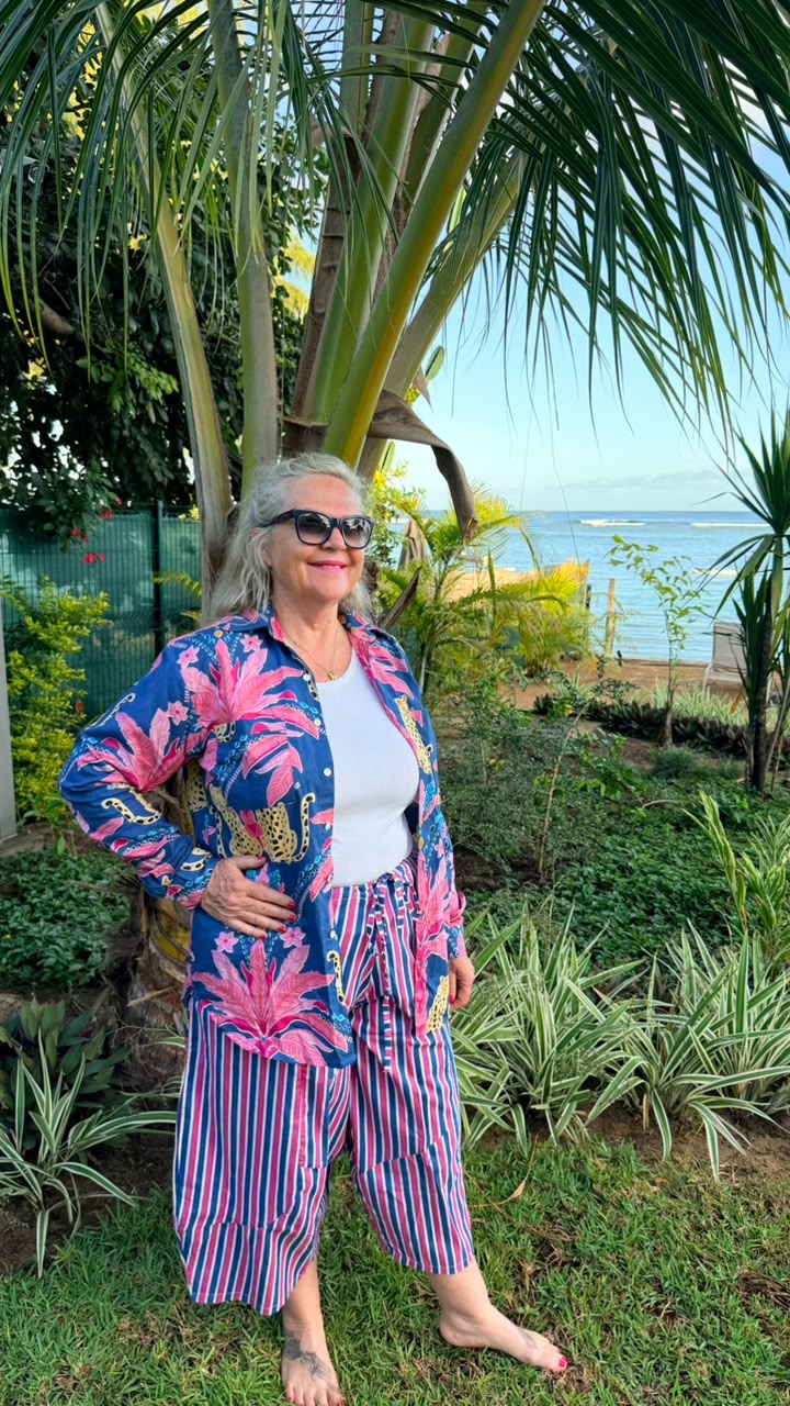 A woman wearing a vibrant tropical print cardigan and striped pants stands outdoors in a garden setting with palm trees and a view of the ocean in the background.