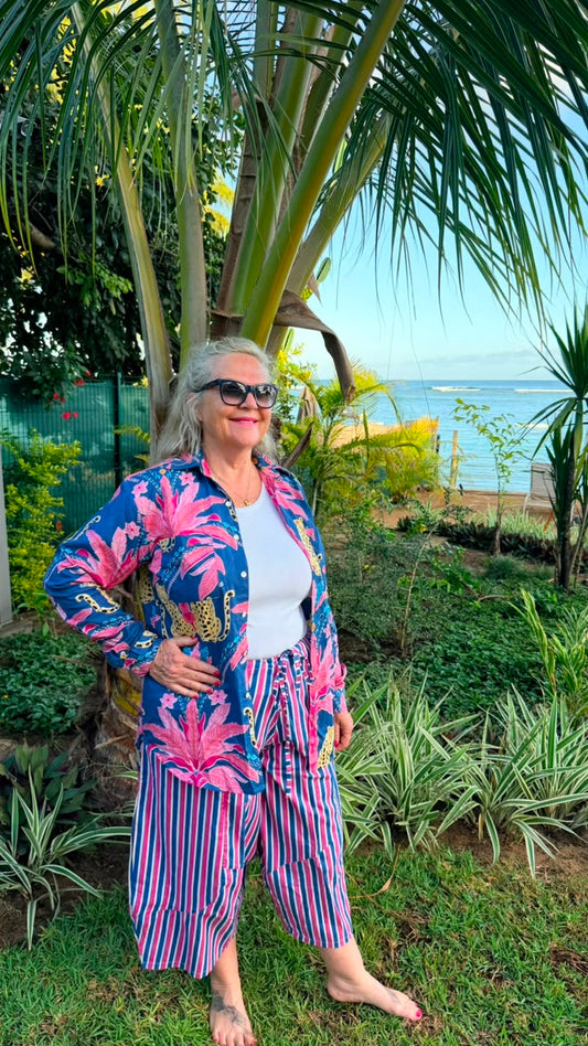 A woman wearing a vibrant tropical print cardigan and striped pants stands outdoors in a garden setting with palm trees and a view of the ocean in the background.