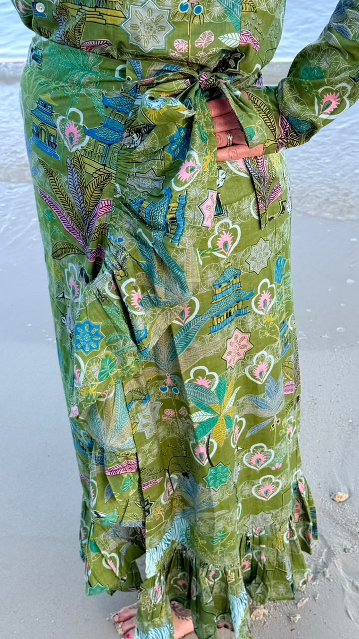 A close-up of a person wearing a green patterned wrap skirt with a tie waist, standing on a sandy beach with water in the background.