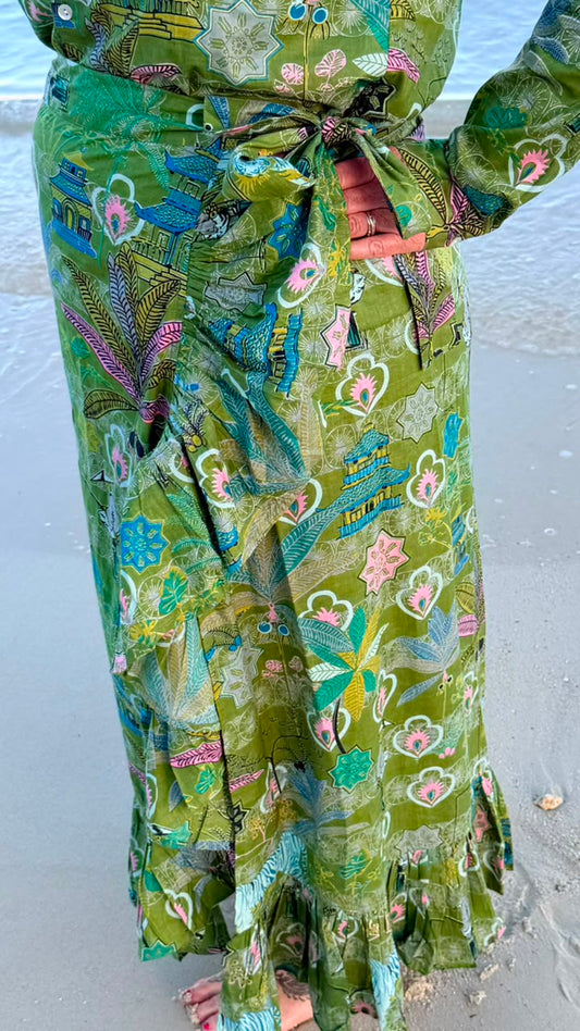 A close-up of a person wearing a green patterned wrap skirt with a tie waist, standing on a sandy beach with water in the background.