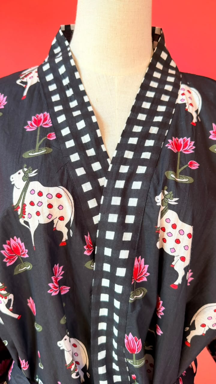 A black kimono-style robe with a white and black checkered border and printed motifs of white cows with red spots, pink lotus flowers, and green leaves, displayed on a mannequin against a red background.