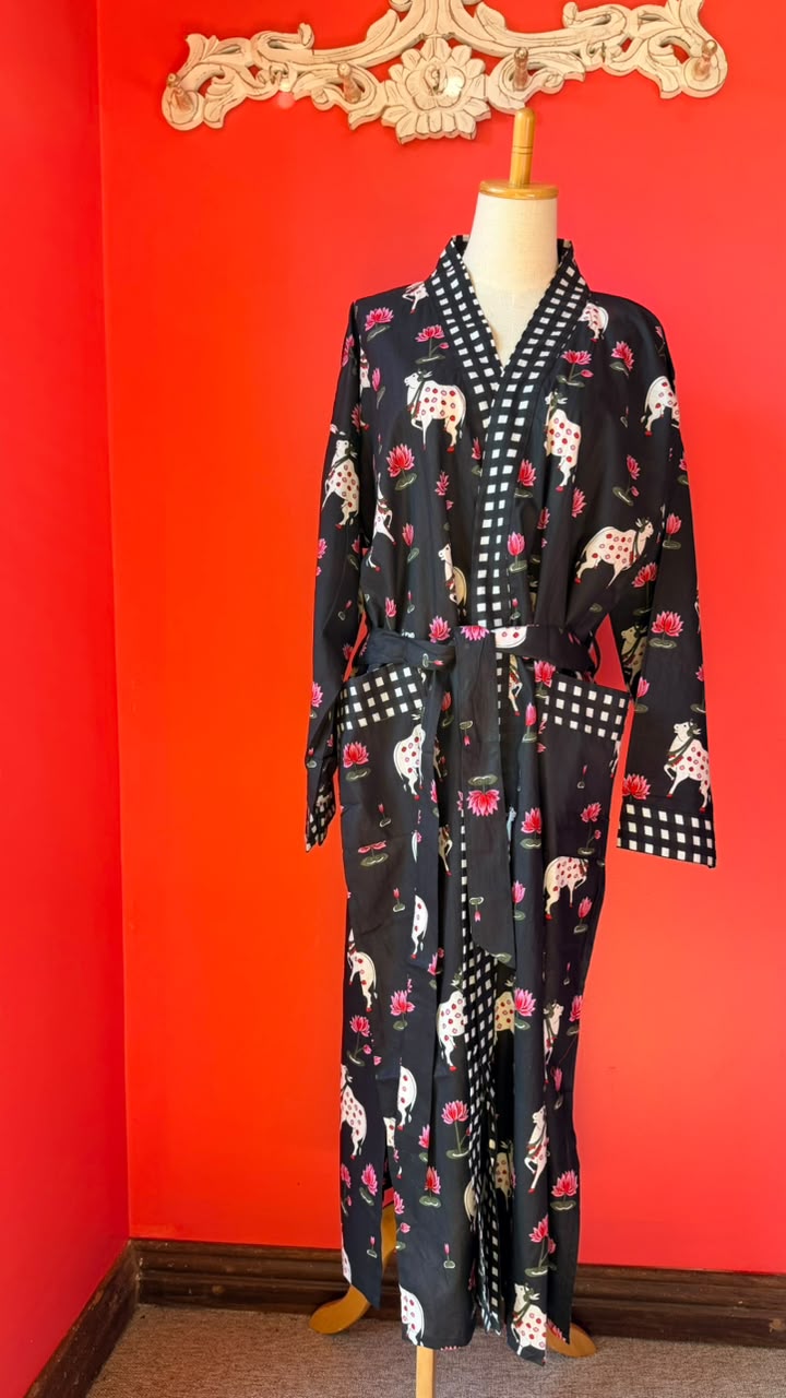 A long black robe with a white and red floral print, displayed on a mannequin against a bright red wall. The robe features a white and black checkered collar, cuffs, and waist tie, and is decorated with illustrations of dogs and roses.