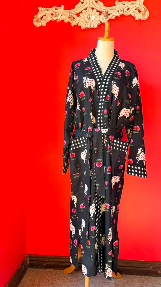A long black robe with a white and red floral print, displayed on a mannequin against a bright red wall. The robe features a white and black checkered collar, cuffs, and waist tie, and is decorated with illustrations of dogs and roses.