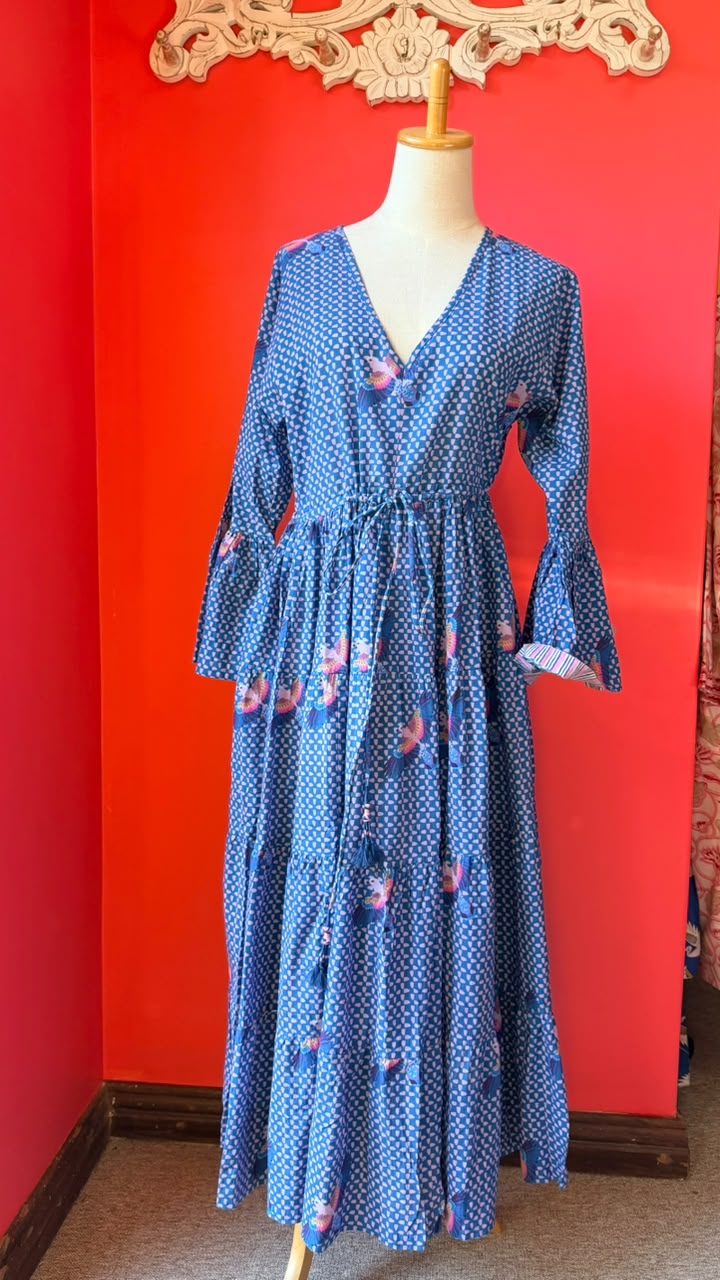 A blue long-sleeve midi dress with a white polka dot pattern and pink floral accents, displayed on a mannequin against a bright red wall.
